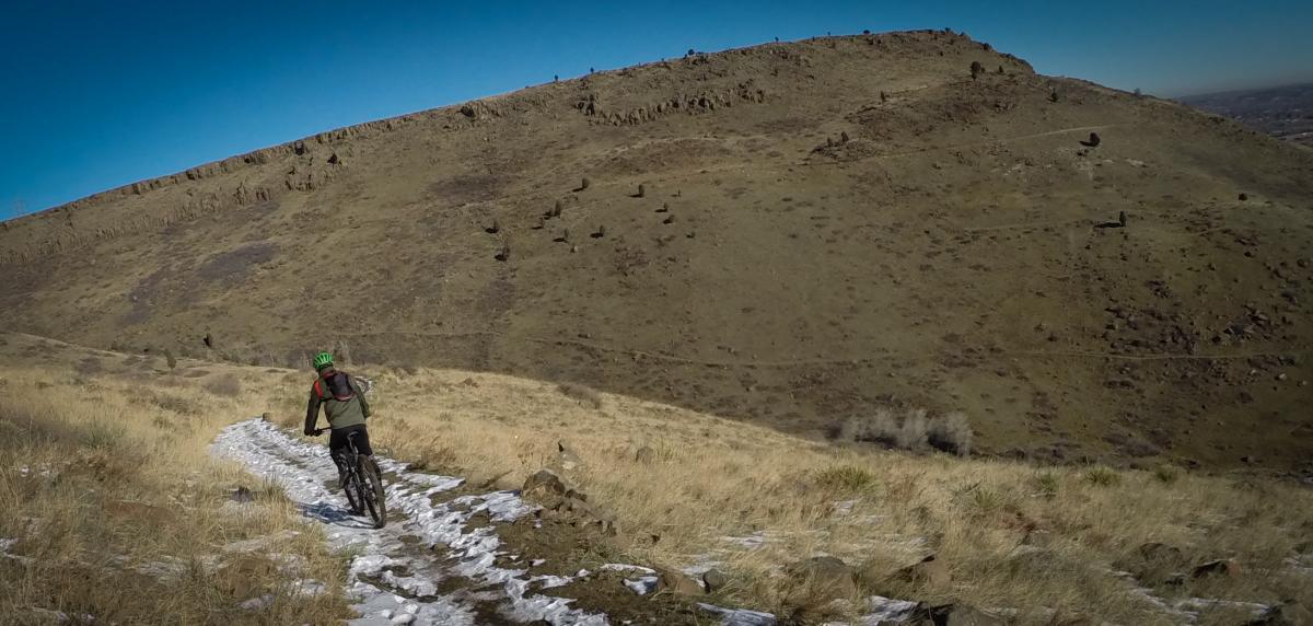 A mountain biker riding along a narrow, snow-dusted path on a hillside, surrounded by dry grass and rocky terrain, with a clear blue sky overhead. North Table Mountain mountain bike trail.
