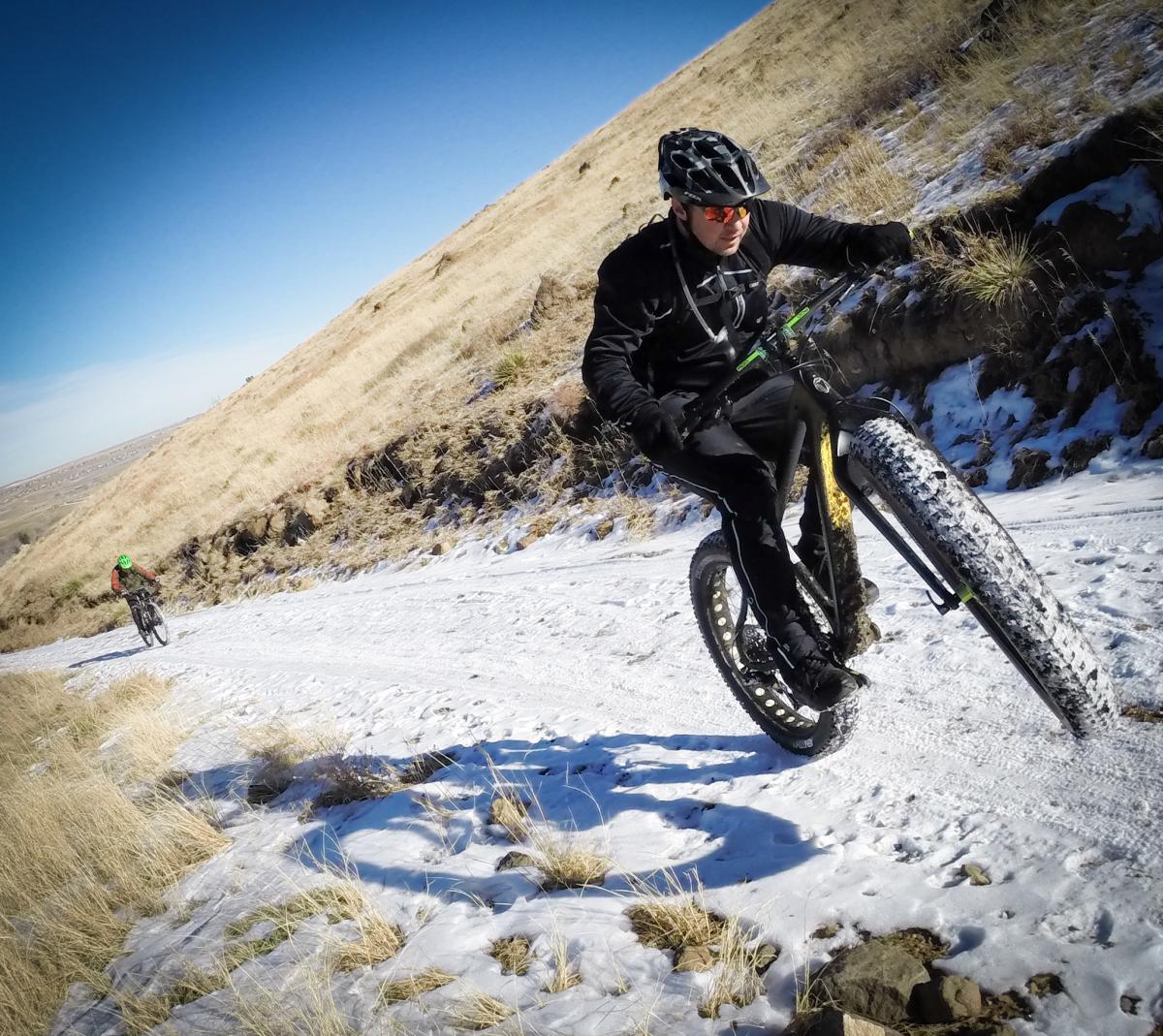 A person riding a fat tire bike on a snowy trail, with another cyclist in the background. The setting features dry grass and a clear blue sky, with a hillside in the distance. The cyclist in the foreground has a focused expression, wearing a helmet and sunglasses, as they perform a wheelie on the bike. North Table Mountain mountain bike trail.