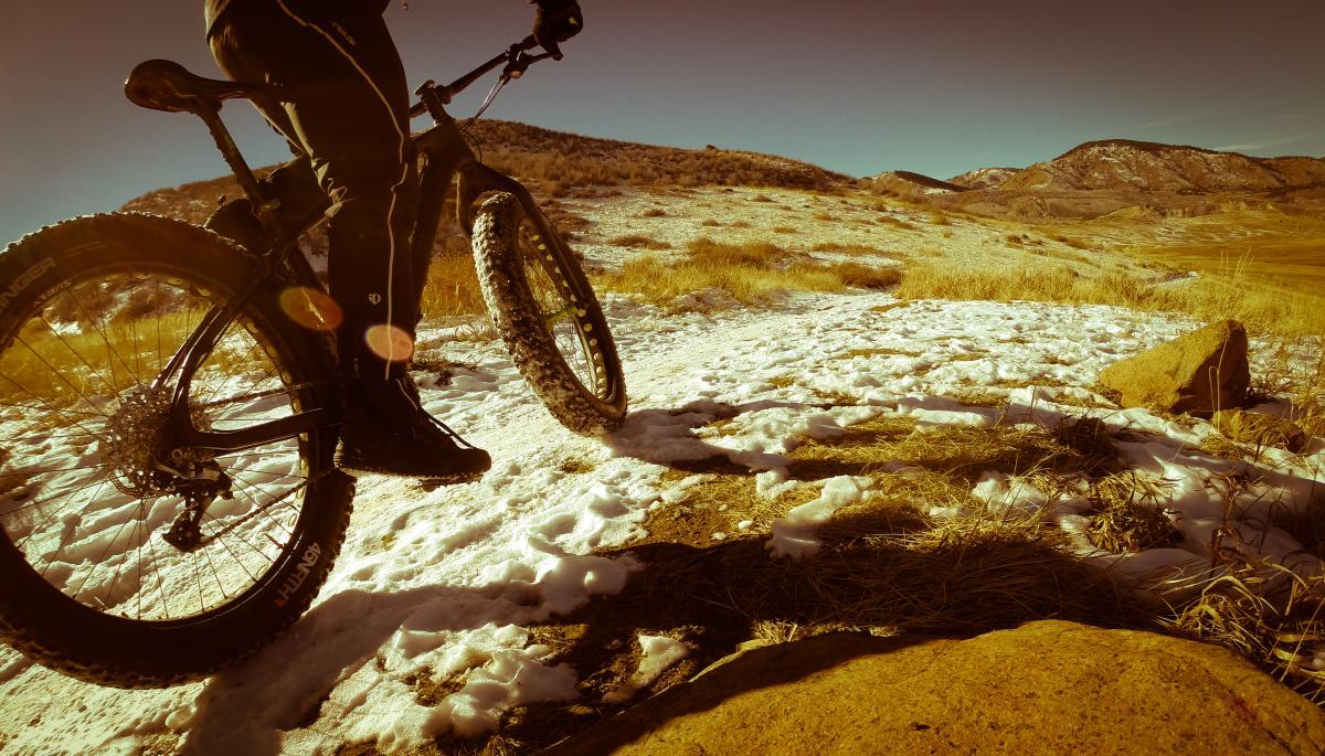 A close-up view of a fat bike parked on a snowy trail, with a person in black cycling gear standing next to it. The landscape features rolling hills and rocky terrain, illuminated by warm sunlight, creating a serene outdoor atmosphere. North Table Mountain mountain bike trail.