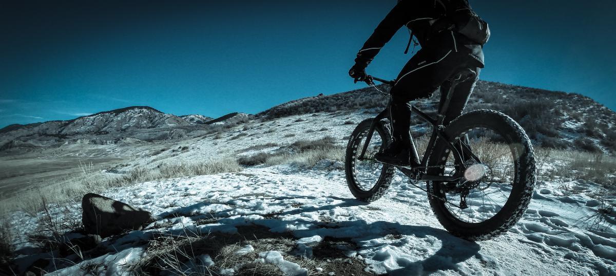 A cyclist riding a fat bike on a snowy, mountainous trail under a clear blue sky. The landscape features rolling hills and patches of grass peeking through the snow. The cyclist is dressed in winter gear, showcasing the active outdoor lifestyle. North Table Mountain mountain bike trail.