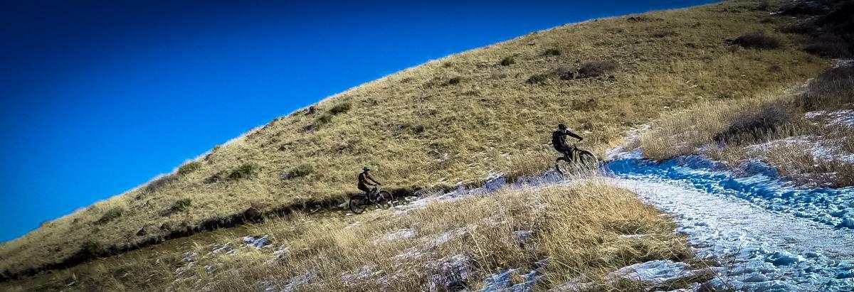 Two mountain bikers riding on a narrow trail through tall grass and snow, with a clear blue sky above. One cyclist is navigating a slight incline while the other is further down the trail. North Table Mountain mountain bike trail.