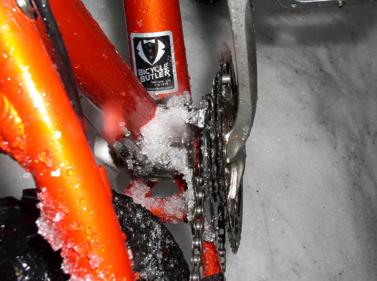 Close-up of an orange bicycle frame covered in snow and ice, focusing on the chain, gears, and a sticker that reads "Bicycle Butler." The background consists of a snowy surface.