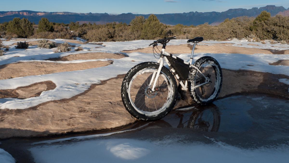 A fat tire bicycle parked on rocky terrain covered in snow and ice, with a scenic backdrop of mountains and trees under a clear blue sky. Gooseberry Mesa mountain bike trail.