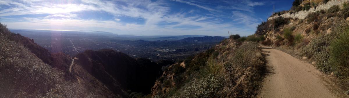 A panoramic view of a mountainous landscape with a dirt trail winding through lush vegetation, overlooking a valley below and distant mountains. The sky is partially cloudy, with sunlight illuminating the scene. Mount Lowe Railway mountain bike trail.