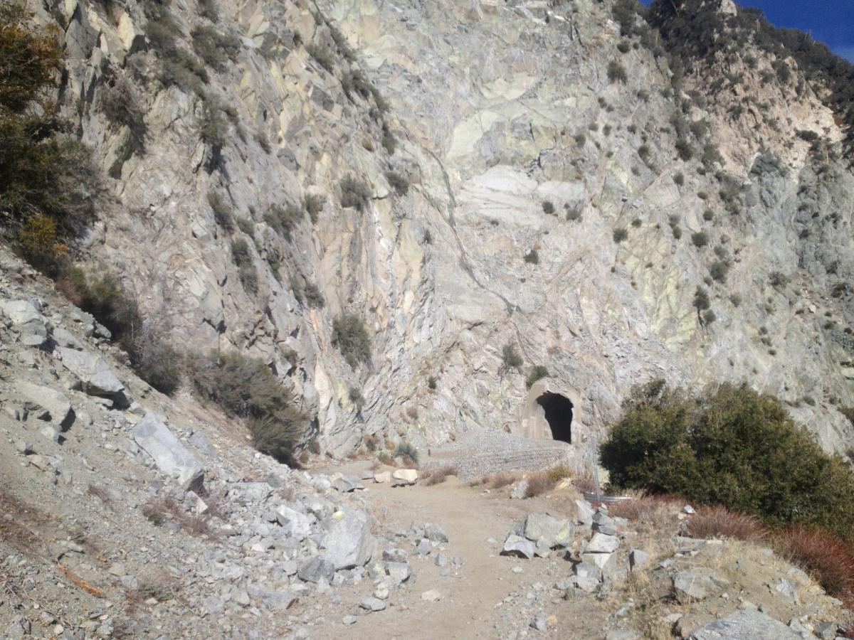 A rocky mountainside featuring a tunnel entrance carved into the hillside, with a dirt path leading towards it. Sparse vegetation surrounds the area, and the rugged terrain is dotted with small rocks and boulders. Mount Lowe Railway mountain bike trail.