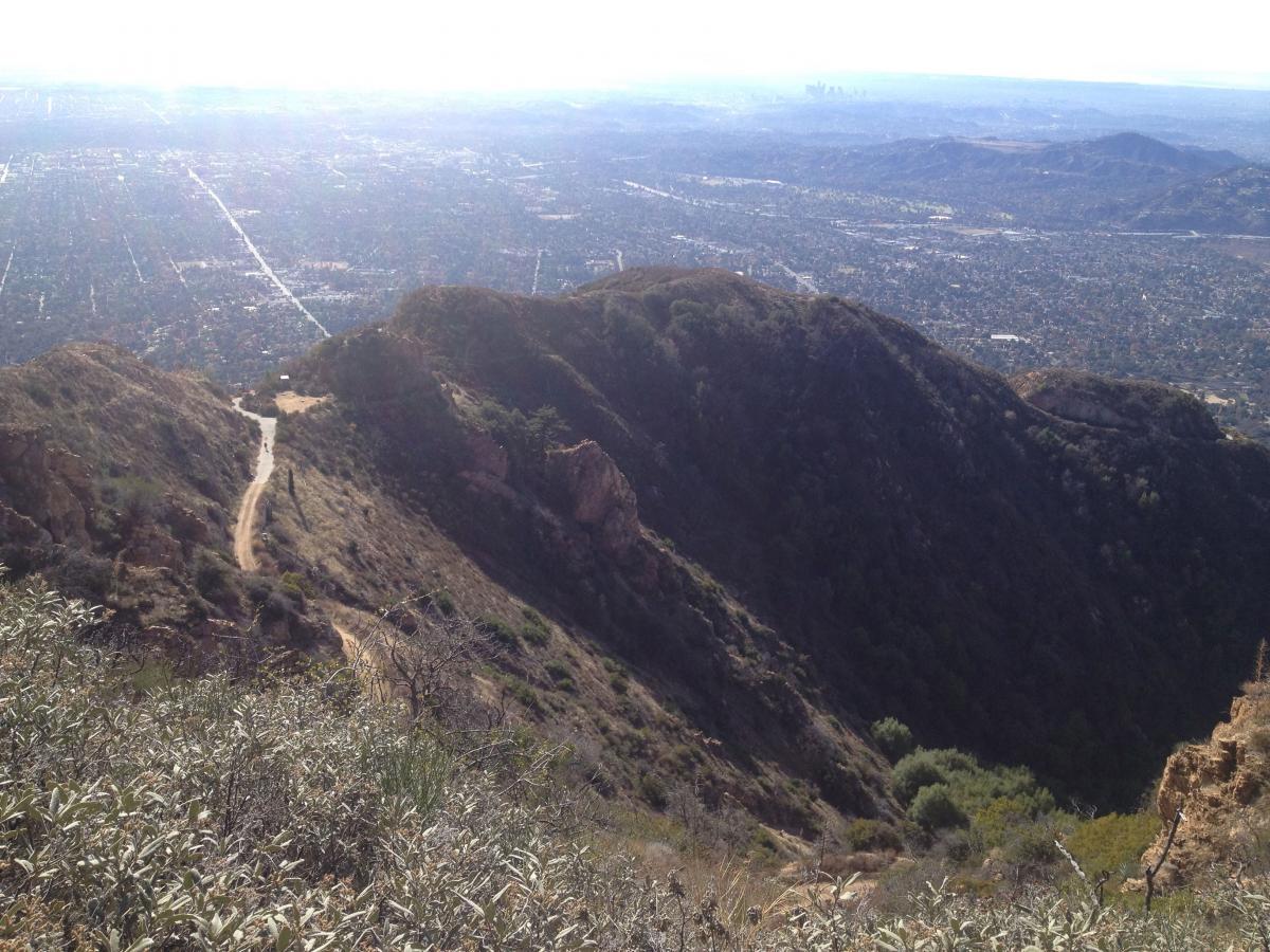 A panoramic view from a hilltop overlooking the sprawling landscape of a city below, with winding paths, hilly terrain, and distant mountains. The scene captures a mix of urban development and natural greenery, under a bright sky, hinting at a sunny day. Mount Lowe Railway mountain bike trail.