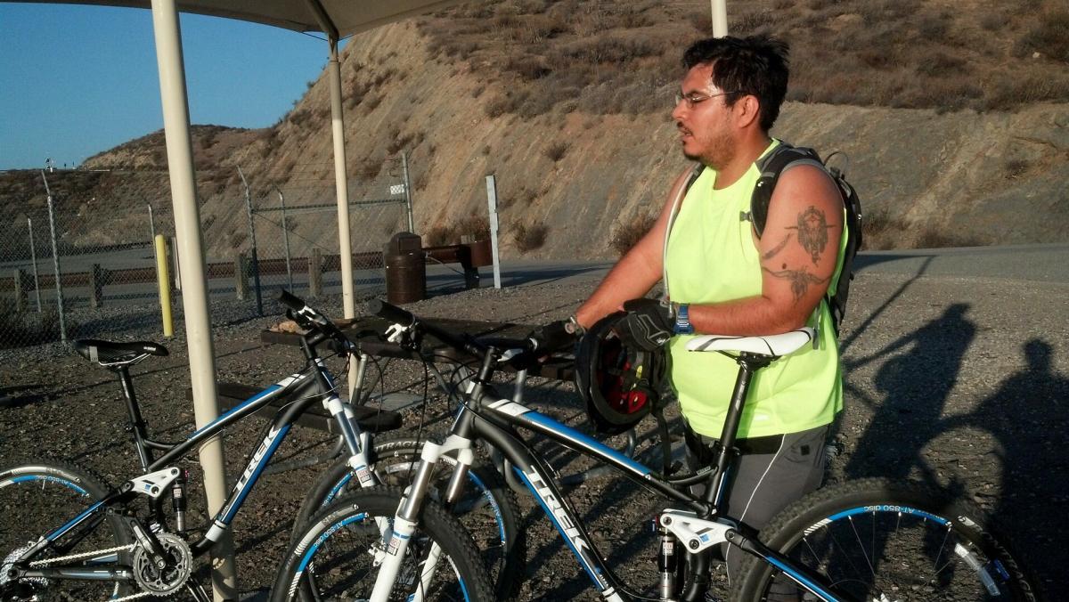 Trek Fuel EX 5: A man wearing a bright yellow sleeveless shirt and gloves stands next to two mountain bikes under a shaded area. He appears to be preparing for a ride, holding a helmet in one hand while looking off to the side. The background features a rocky hillside and a dirt path, with a chain-link fence visible in the distance. The lighting suggests a late afternoon setting.