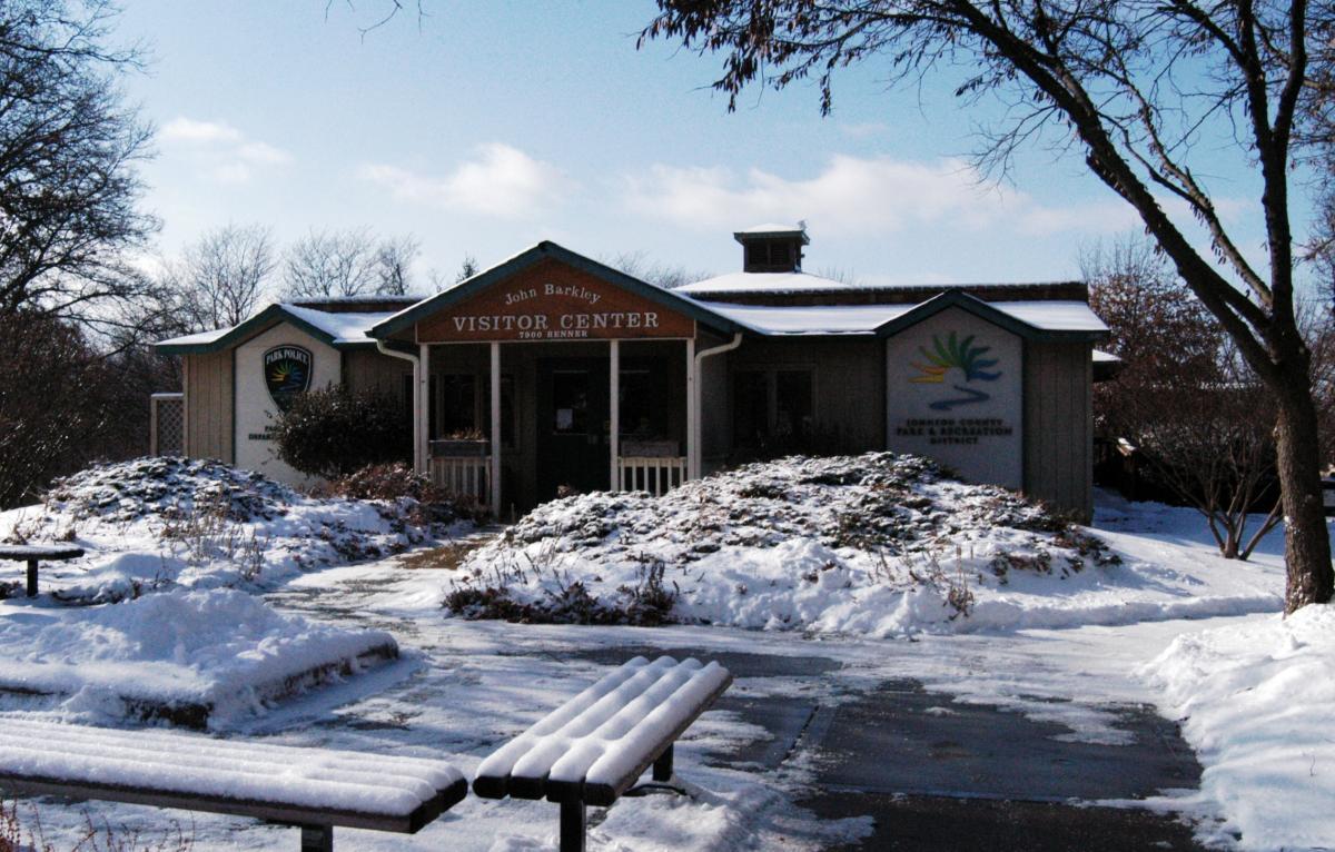 A cozy visitor center covered in snow, featuring a sign that reads "John Barkley Visitor Center." The building is surrounded by snowy landscapes and bare trees, with benches in front. A clear blue sky peeks through fluffy clouds above. Shawnee Mission Park mountain bike trail.