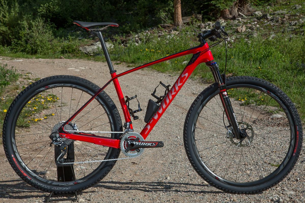 Specialized STUMPJUMPER EXPERT CARBON HT: A red S-Works mountain bike parked on a gravel path surrounded by greenery. The bike features a sleek frame design, thick tires, and two water bottle holders, with a clear view of the gear mechanism.
