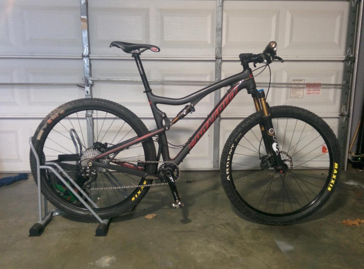 Santa Cruz Tallboy Carbon: A mountain bike with a black and red frame is placed on a bike stand in a garage. The bike features thick tires, a front suspension fork, and a single chainring setup. The background includes a closed garage door and some scattered items on the floor.