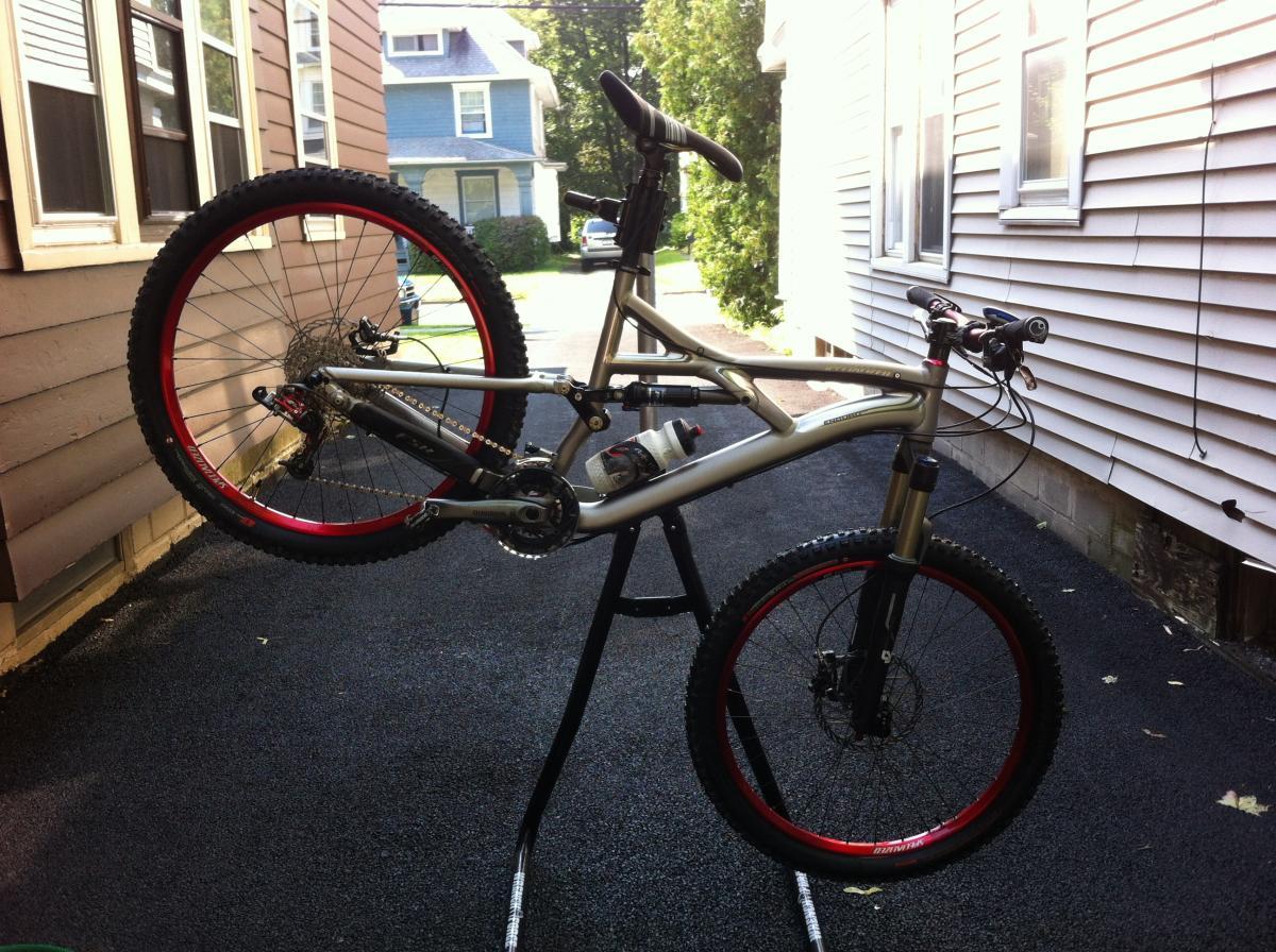 Specialized Enduro SL Expert: A mountain bike displayed on a repair stand in a driveway, showcasing a silver frame and red wheels. The bike is positioned upright, highlighting its components, including the handlebars and suspension fork. In the background, a residential area with houses and greenery is visible.
