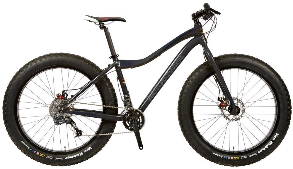 Motobecane Boris: A side view of a fat tire bike with oversized tires designed for off-road terrain. The bike features a dark frame, disc brakes, and a single gear setup, showcasing its robust design suitable for rough trails and snowy conditions. The tires are labeled "Vee Rubber," emphasizing their durability and grip.