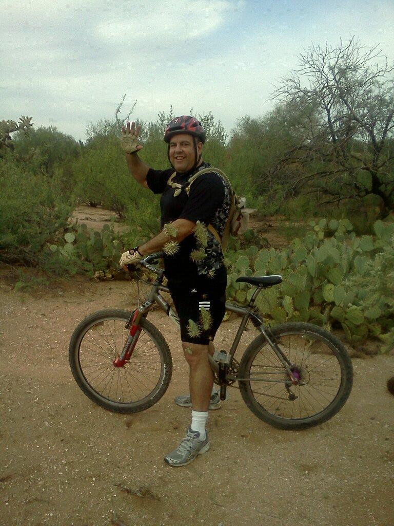 A person in athletic gear, wearing a helmet and smiling, stands next to a mountain bike on a dirt path surrounded by desert vegetation, including cacti. They are raising one hand in a friendly wave. Fantasy Island mountain bike trail.