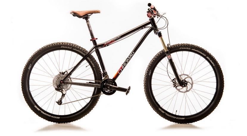 Chromag Rootdown: A black mountain bike with a sleek frame and brown leather saddle, featuring wide, knobby tires for off-road traction. The bike is positioned upright against a plain white background, showcasing its components such as the front suspension fork and disc brakes.