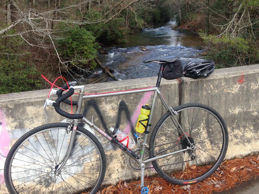 Club Roost Cross Terra Tire: A silver bicycle with a black helmet and water bottle rests against a concrete barrier overlooking a flowing creek. Surrounding the creek are trees with sparse foliage, indicating a late winter or early spring setting. The barrier features some graffiti, adding a splash of color to the natural landscape.