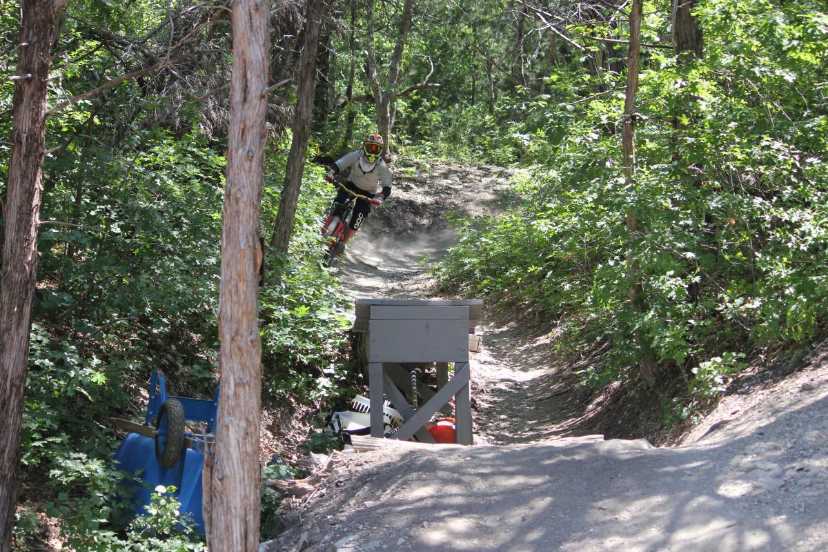 A mountain biker rides a dirt trail through a forested area, kicking up dust as they navigate a jump over a wooden ramp. Surrounding vegetation includes green shrubs and trees, creating a natural, outdoor setting. Big Cedar Wilderness Trails mountain bike trail.