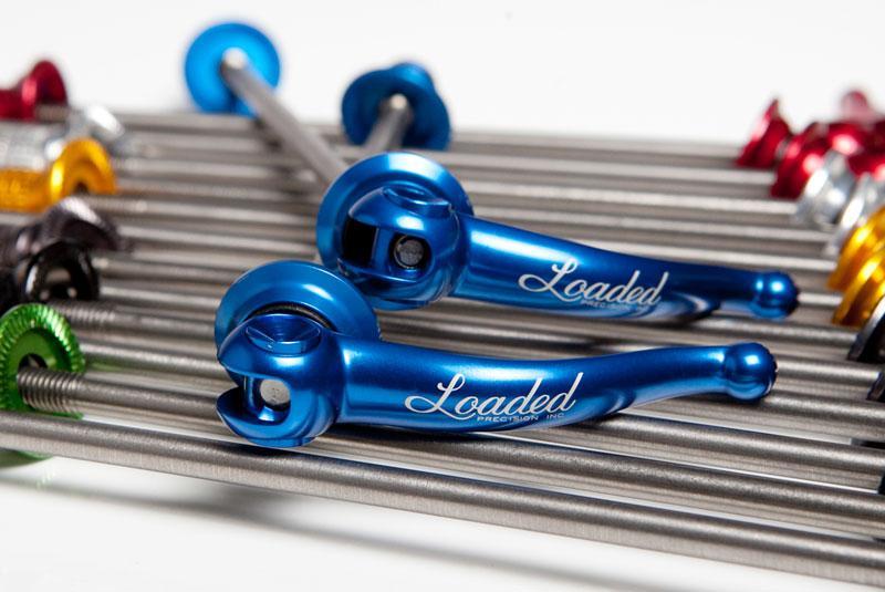 Loaded Precision X-lite Titanium Race Day: Two blue precision components labeled "Loaded" positioned on a metallic background with various colored accessories, such as red, green, silver, and yellow, lined up in an organized manner.