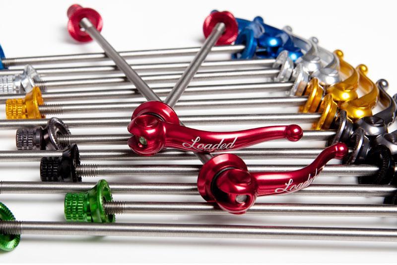 Loaded Precision X-lite Titanium Race Day: A close-up of various bicycle quick-release levers arranged in a dynamic layout. The levers feature different colors, including red, blue, gold, and green, and are made of metallic materials. Some levers have the word "Loaded" engraved on them, highlighting their brand. The background showcases a clean white surface, emphasizing the vivid colors and details of the levers.