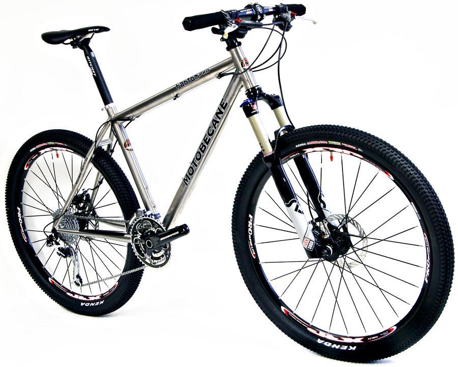 Motobecane Fantom PRO Titanium: Alt text: A silver mountain bike with a lightweight frame, featuring thick black tires and a front suspension. The bike is displayed against a white background, showcasing its sleek design and components. The brand name "Motobecane" is visible on the frame.