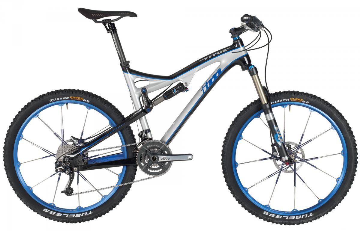 Titus FTM Carbon: Image of a sleek mountain bike featuring a silver and black frame with blue accents. The bike has wide, rugged tires labeled "Rubber Queen" and disc brakes, making it suitable for off-road terrain. The design includes a prominent suspension system and a modern gear setup.