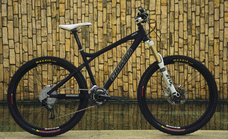 Chumba CHUMBA HX1: A black mountain bike with a white seat, featuring wide tires and a sleek frame, positioned against a textured stone wall with a reflective surface in front. The bike has a front suspension fork and is equipped with high-quality components suitable for off-road cycling.