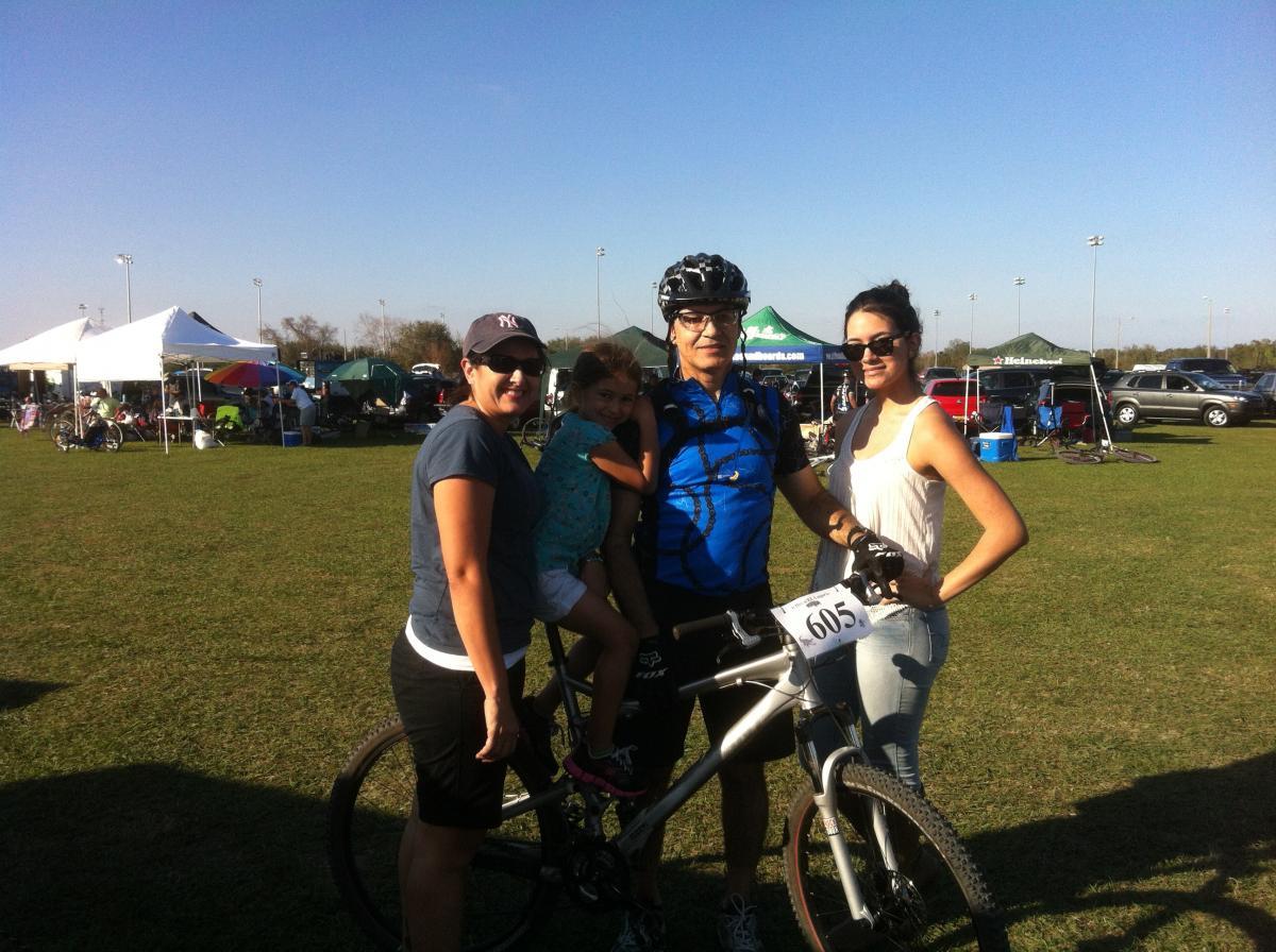 Giant Trance X3: A group of four individuals poses together outdoors, showing a sunny day at a cycling event. One adult holds a mountain bike, while a young girl sits on their hip. The other two adults stand beside them, smiling. In the background, several tents and vehicles are visible on a grassy field.