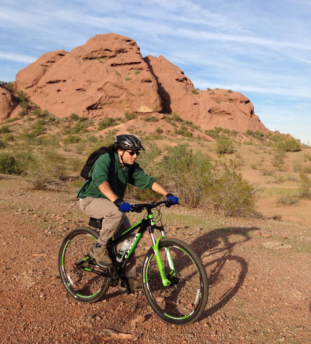 Trek X-Caliber: A person in a green long-sleeve shirt and a black helmet rides a mountain bike on a rocky trail, with reddish rocky hills and sparse vegetation in the background under a blue sky.