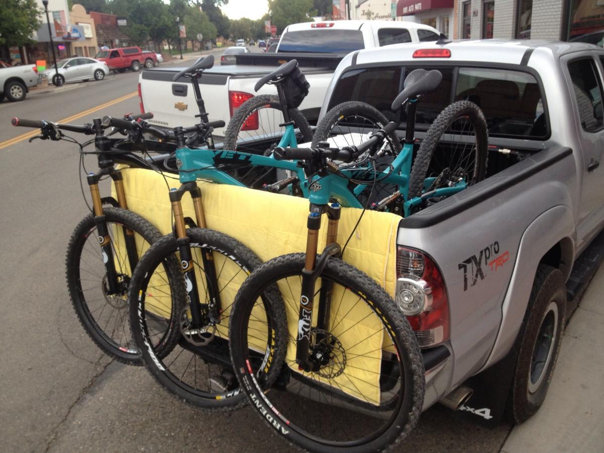Yeti SB95: A silver pickup truck with mountain bikes loaded in the truck bed, featuring three bikes with yellow padding for protection. The background includes a street scene with parked cars and storefronts.