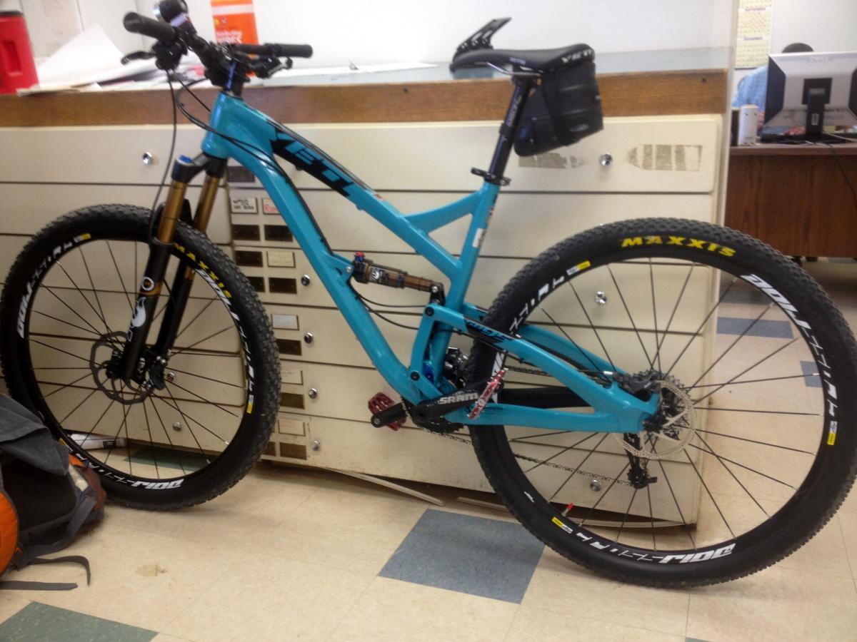 Yeti SB95: A turquoise mountain bike leaning against a filing cabinet, featuring large, knobby tires and a front suspension fork. The bike has a sleek frame and a black saddle with a small accessory pouch attached. In the background, there is a glimpse of an office setting, including a desk and computer.