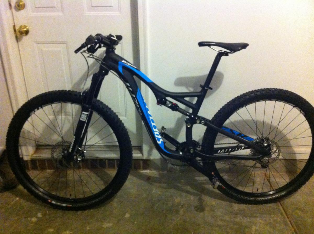 Specialized Stumpjumper FSR Comp 29: A black and blue mountain bike leaning against a wall, with thick tires and a front suspension fork, positioned in a garage space. The door and floor are partially visible in the background.