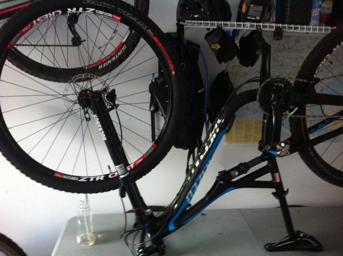 Specialized Stumpjumper FSR Comp 29: A mountain bike displayed vertically on a wall rack, with one wheel removed and resting on a table below. The bike features a black and blue frame, visible components include the drivetrain and suspension system. Above, additional bicycle wheels and helmets are hanging on hooks. A clear bottle is positioned on the table. The background contains a map and some bags.