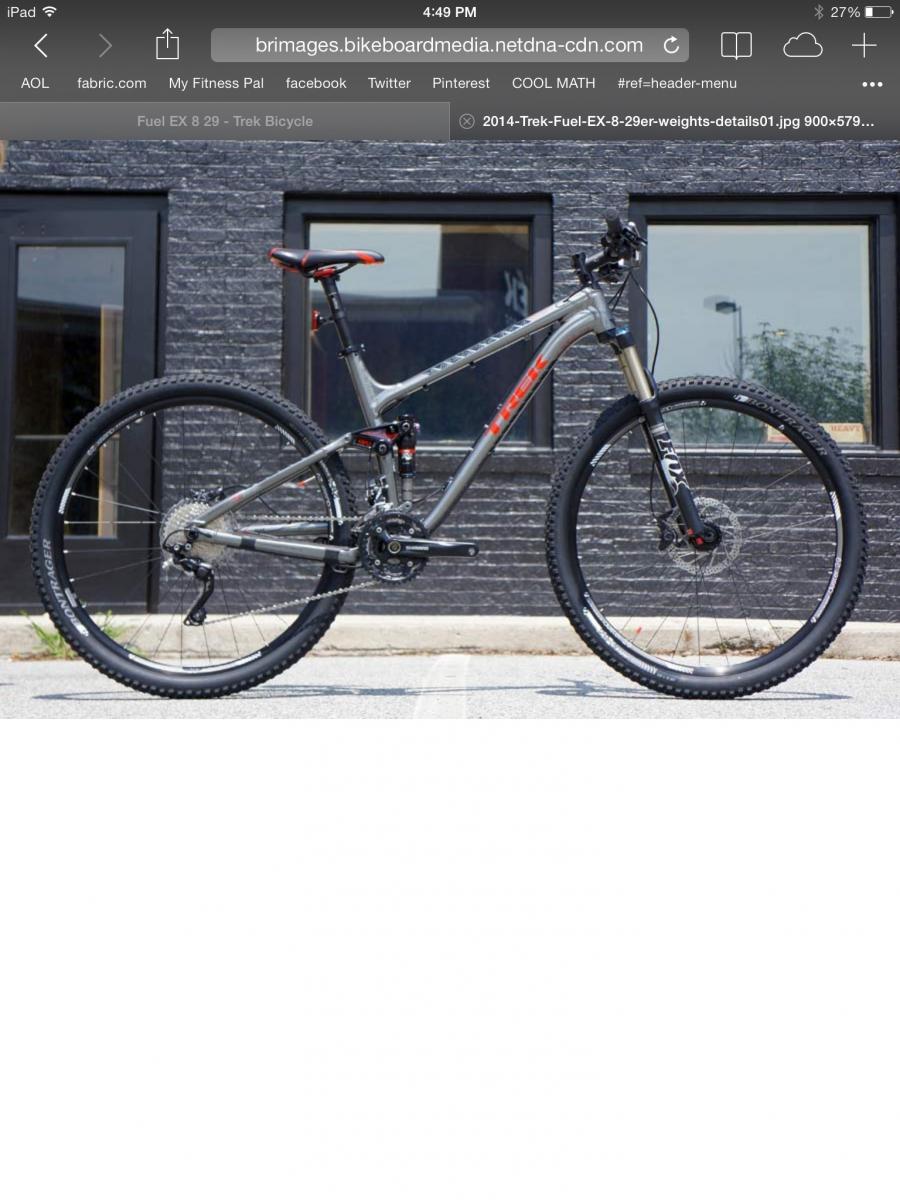 Trek Fuel EX 8: A Trek Fuel EX 8 29 mountain bike featuring a silver and red design, standing on a paved surface in front of a textured dark wall. The bike has thick tires, a prominent suspension fork, and a sleek, lightweight frame.