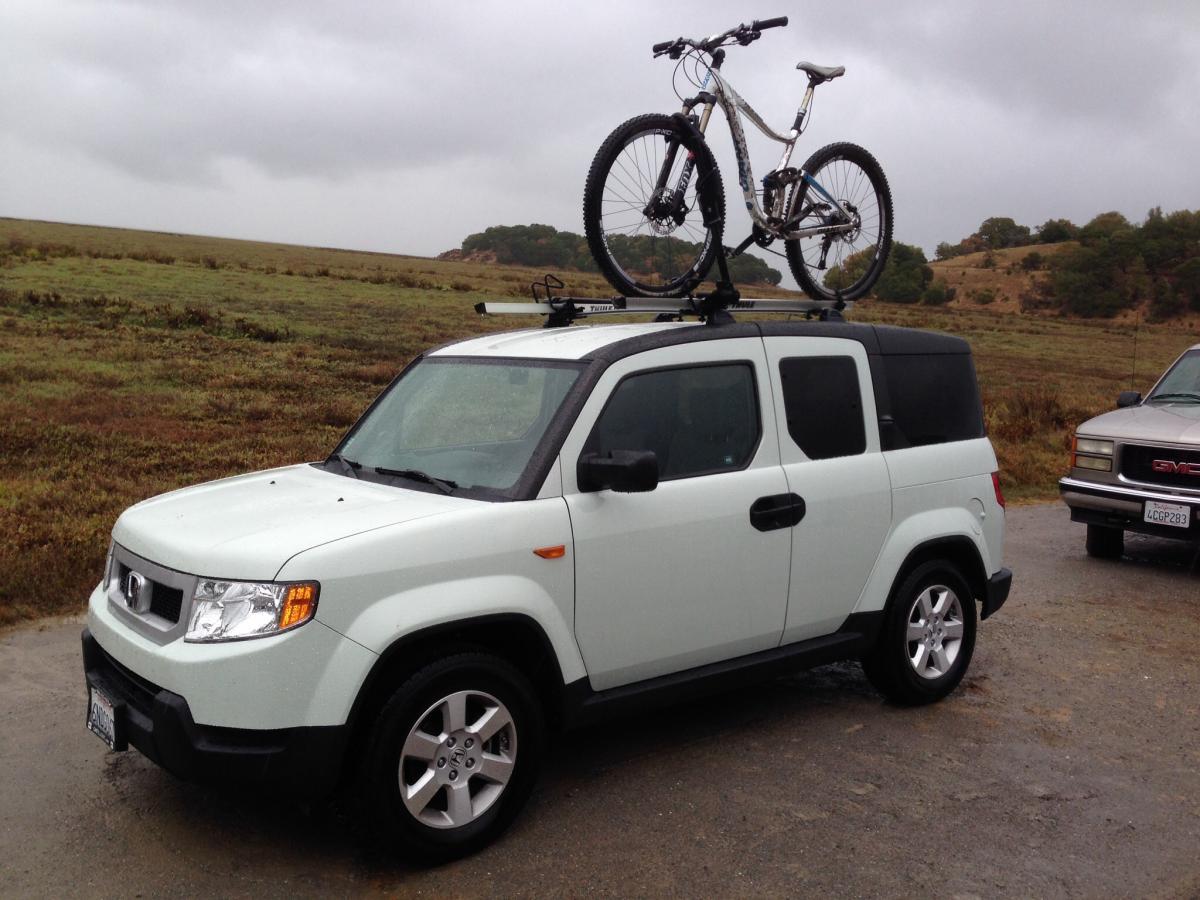 A light-colored SUV parked on a gravel surface, with a mountain bike mounted on its roof rack. The background features a grassy field and rolling hills under a cloudy sky. China Camp mountain bike trail.