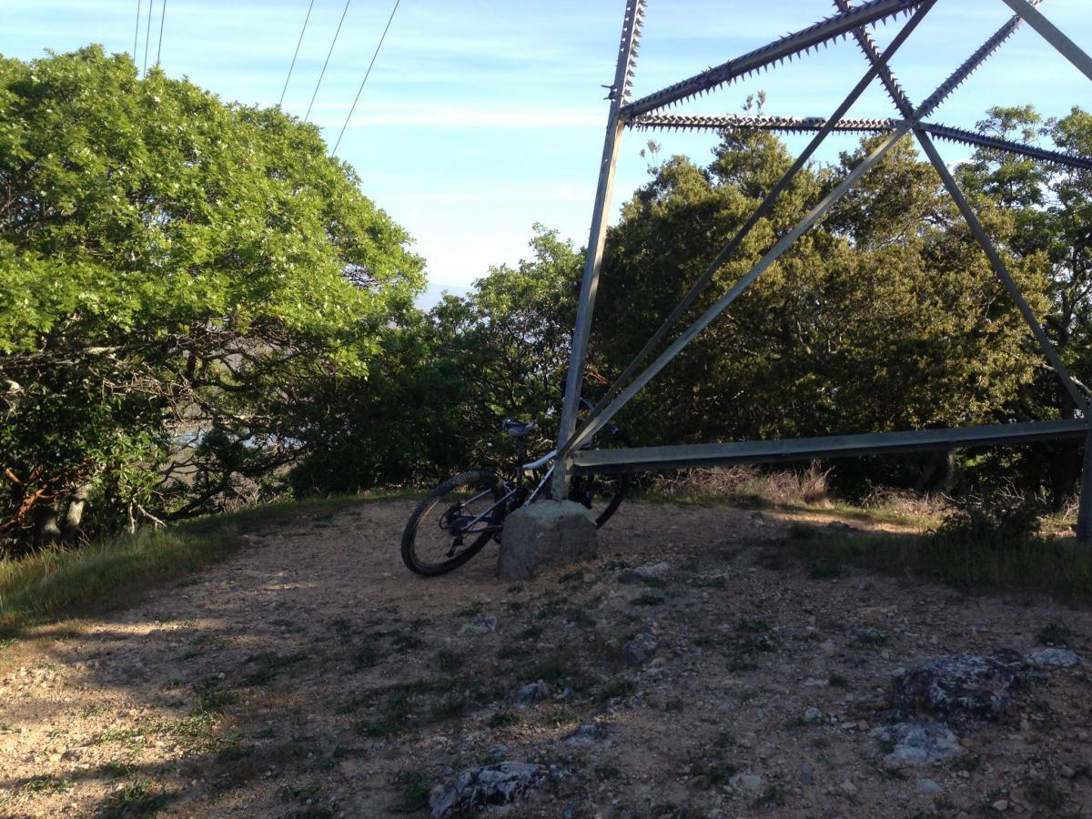 A mountain bike leaning against a concrete block near a power transmission structure, surrounded by green trees and a dirt path. China Camp mountain bike trail.