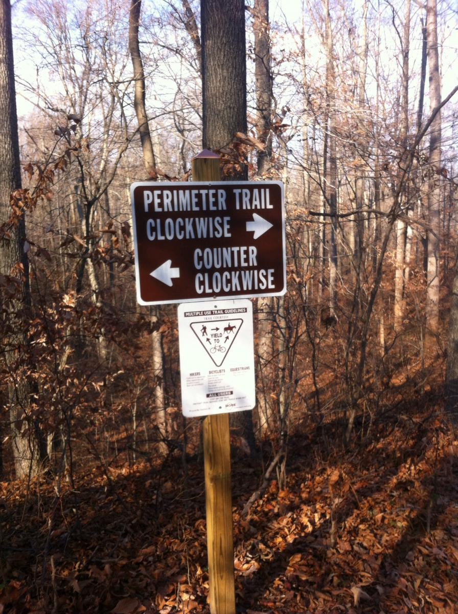 Brown trail sign indicating directions for the Perimeter Trail, with arrows pointing "Clockwise" and "Counter Clockwise." A secondary sign below provides guidelines for yielding to hikers, cyclists, and equestrians. The background features a wooded area with bare trees and fallen leaves. Rosaryville State Park mountain bike trail.