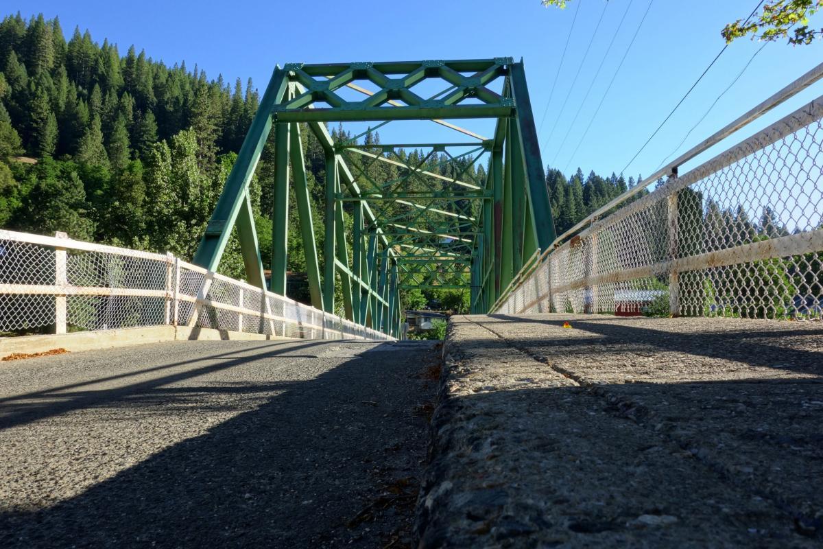 A low-angle view of a green metal truss bridge, showcasing its structure and extending over a road. The bridge is flanked by a white chain-link fence and surrounded by lush green trees and forested hills under a clear blue sky. Downieville Downhill mountain bike trail.