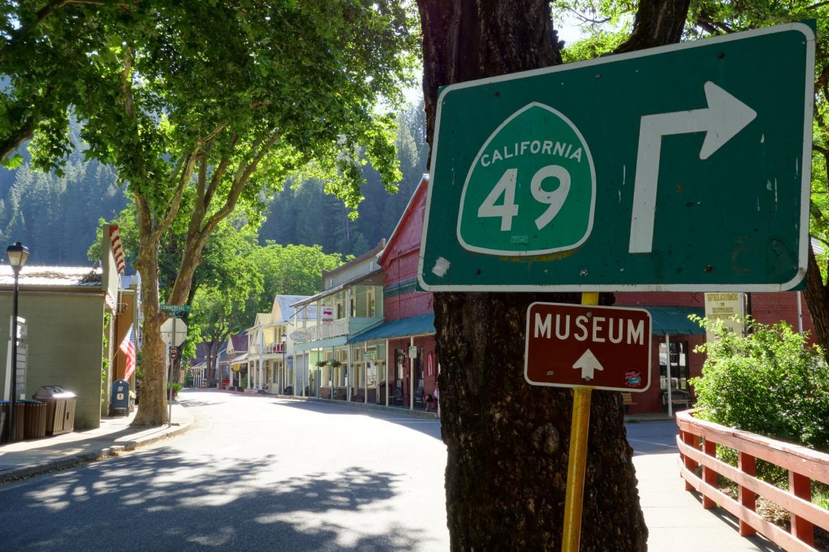 Signposts for California Highway 49 with an arrow indicating a right turn, alongside a directional sign pointing to a museum, set against a backdrop of trees and colorful historic buildings lining a quiet street. Downieville Downhill mountain bike trail.