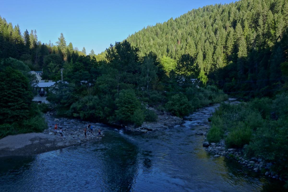 A serene landscape featuring a tranquil river flowing through a forested area, surrounded by lush green trees and a clear blue sky. Small groups of people are seen enjoying the water and rocky riverbank, while a building is nestled among the trees in the background. Downieville Downhill mountain bike trail.