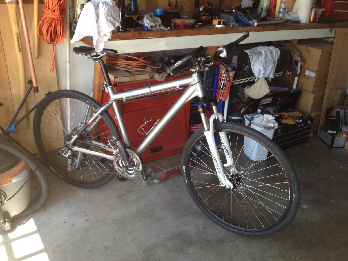 Giant Seek: A silver bicycle leaning against a cluttered garage wall, surrounded by tools and items like a vacuum, a toolbox, and various outdoor equipment. The garage has a mix of organized and disorganized elements, with items scattered on a shelf above the bicycle.