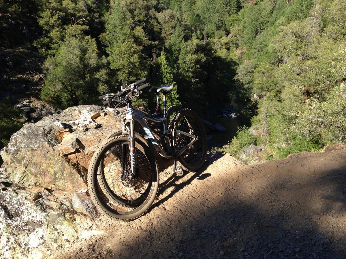 Giant Trance X 29er: A mountain bike resting on rocky terrain, overlooking a lush, green forested area with a stream visible in the background. The scene is illuminated by bright sunlight, showcasing the natural beauty of the outdoors.