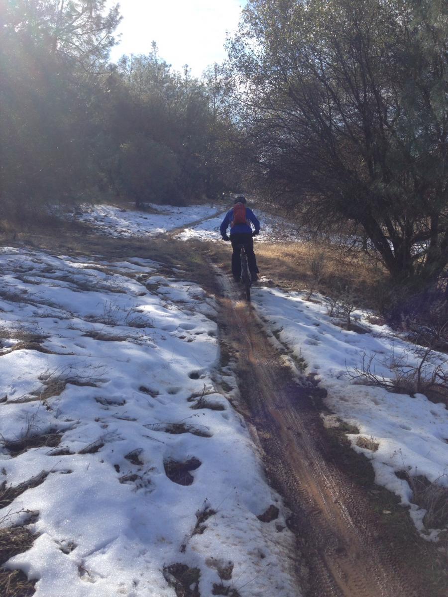 A person riding a mountain bike along a snowy trail surrounded by trees. Sunlight filters through the foliage overhead, illuminating the path that has both snow and patches of bare ground. The rider is seen from behind, wearing a blue jacket and a red backpack. Clementine / Forresthill Connector Trail mountain bike trail.