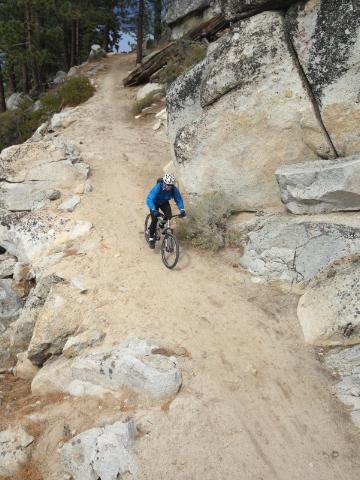 A mountain biker navigating a rocky dirt trail surrounded by trees and large boulders. Flume Trail mountain bike trail.