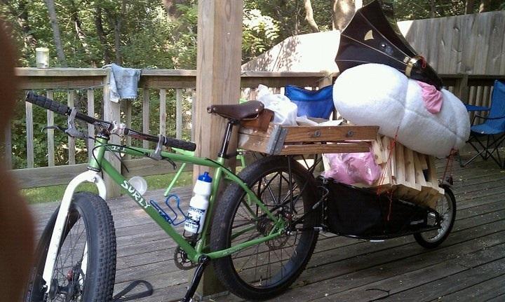 Surly 1x1: A green bicycle with a large rear cargo attachment is parked on a wooden deck. The cargo area is filled with various items, including a white cylindrical object, wooden planks, and bundles of fabric. A water bottle is mounted on the bike frame, and chairs can be seen in the background surrounded by trees.