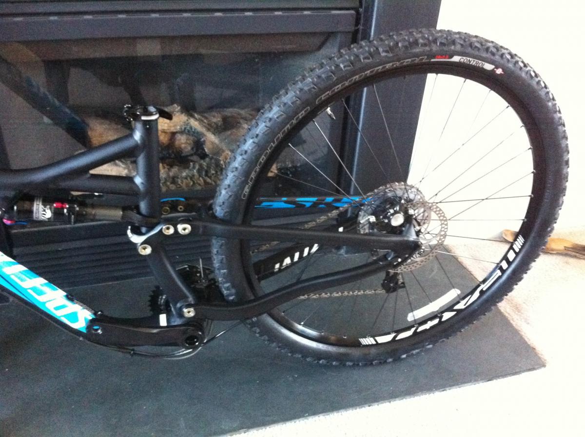 Specialized Ground Control: A close-up view of the rear wheel and frame of a mountain bike, featuring a black frame with blue accents. The background includes a fireplace with visible stones. The bike is situated on a dark surface, highlighting its design and components.