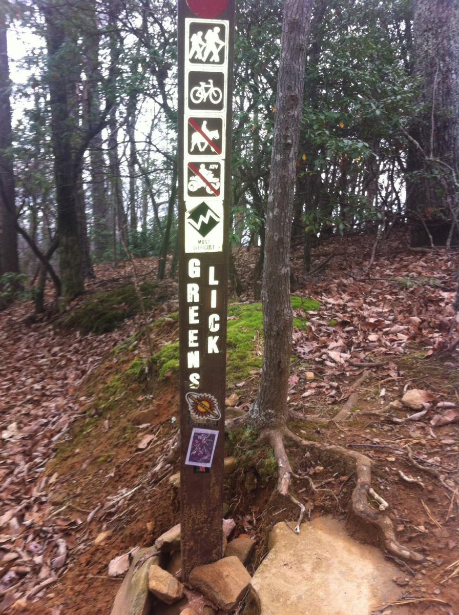 A trail marker sign in a wooded area with icons indicating allowed activities such as hiking and biking, while prohibiting dogs and ATVs. The sign features the name "Greens Lick" prominently, and is surrounded by leaf litter and moss. Various stickers are affixed to the post. Bent Creek mountain bike trail.