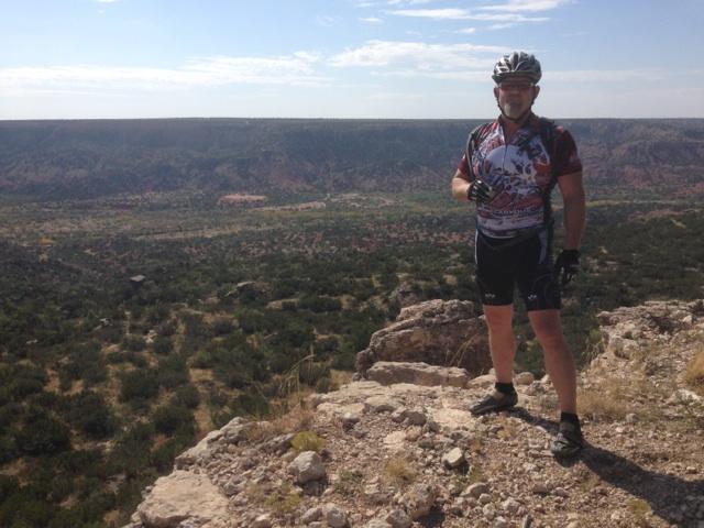 Niner Air 9: A cyclist stands on a rocky outcrop, overlooking a vast canyon landscape. The sky is blue with a few clouds, and the terrain is filled with greenery and distant rock formations. The cyclist is wearing a colorful cycling jersey, shorts, and a helmet, smiling as they enjoy the scenic view.