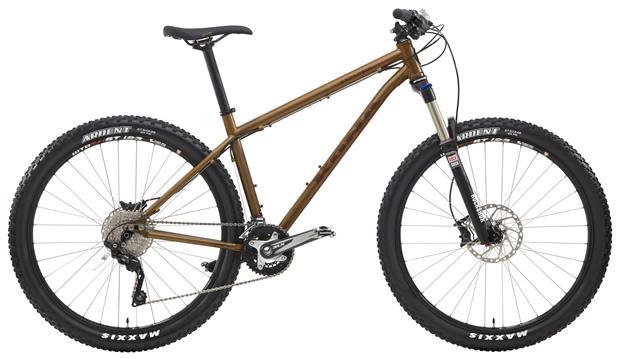 Kona EXPLOSIF: A mountain bike with a brown frame, equipped with thick tires and a front suspension fork. The bike features disc brakes, a gear shifter, and a Shimano drivetrain, showcasing a sturdy design suitable for off-road riding.