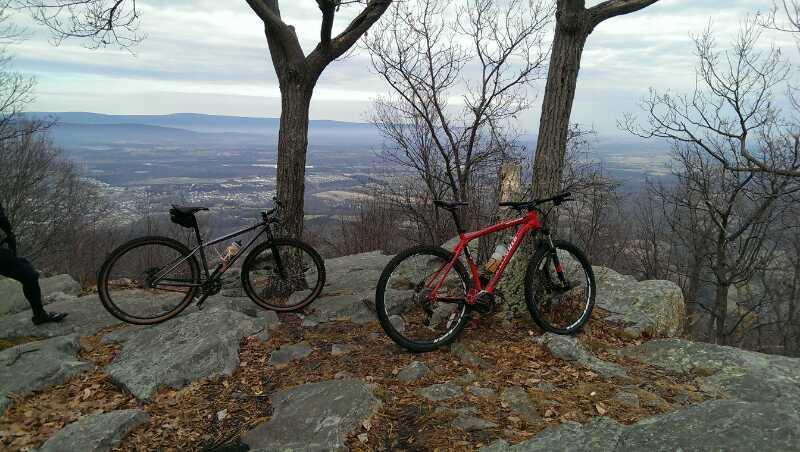 Two mountain bikes, one black and one red, are parked on a rocky outcrop with a scenic view of a valley and distant mountains. The trees are bare, indicating a late autumn or early winter setting, and fallen leaves cover the ground. The sky is cloudy, adding a soft light to the landscape. Elizabeth Furnace mountain bike trail.