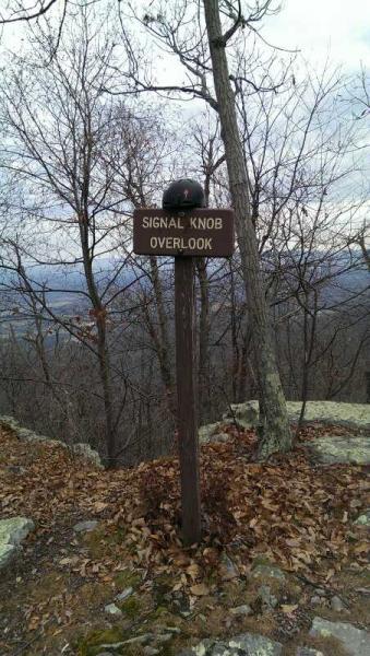 A wooden sign labeled "Signal Knob Overlook" stands on a rocky path surrounded by bare trees, with a cloudy sky overhead. Fallen leaves cover the ground, and the vast landscape is visible in the background. Elizabeth Furnace mountain bike trail.