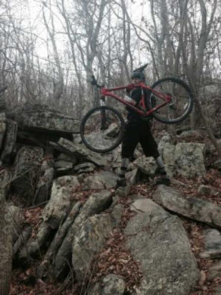 A person in cycling gear stands on rocky terrain, holding a red mountain bike above their head. The background features bare trees and scattered rocks, indicating a challenging outdoor environment. Elizabeth Furnace mountain bike trail.